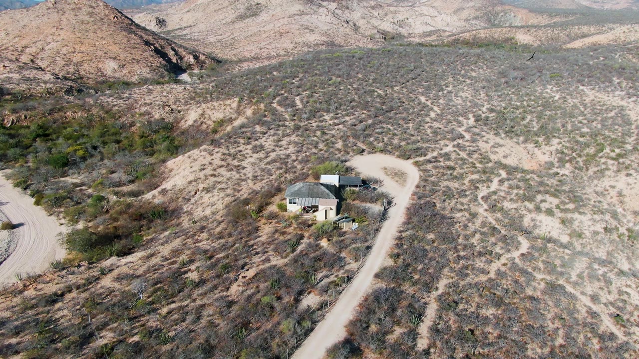 Opening aerial frame over Cabo Pulmo desert terrain, showing a small light-roofed building near a curving dirt road, scrub vegetation, low hills, and no visible people.