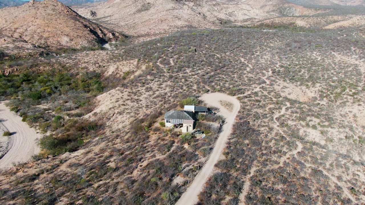 Drone-height view of a remote structure in Cabo Pulmo desert, with scrub-covered slopes, pale tracks, a curving road, distant hills, and no visible people.