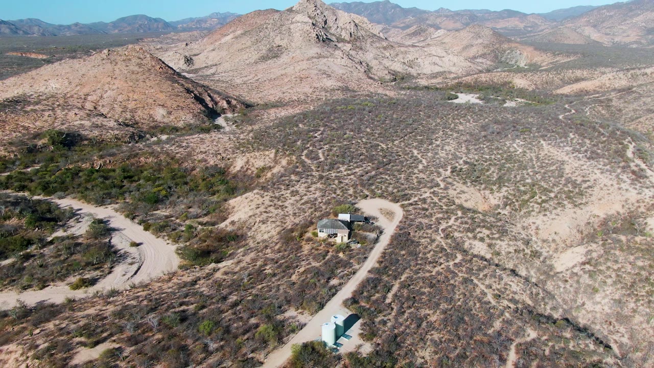 High aerial view over Cabo Pulmo desert terrain with a winding dirt road, small structures near the lower frame, rocky hills, distant mountains, clear sky, and no visible people.