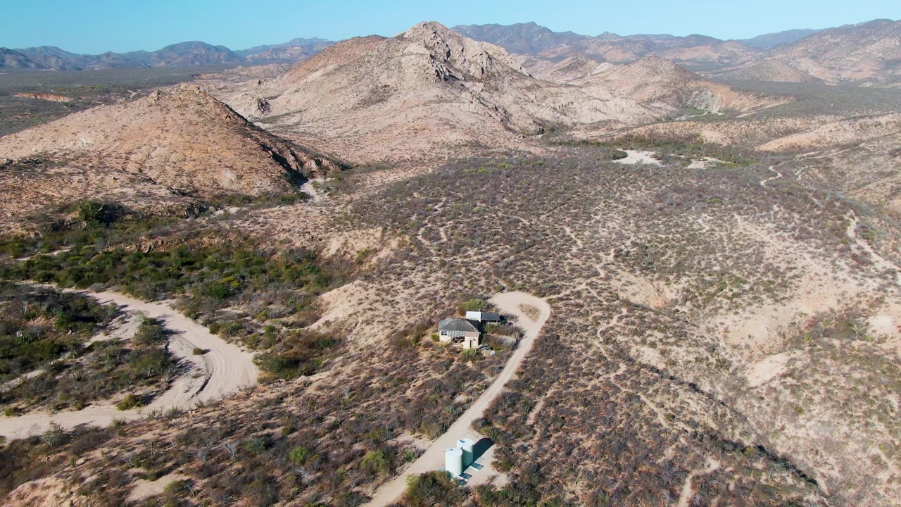 Desert road curves past small buildings below rocky hills and mountains.