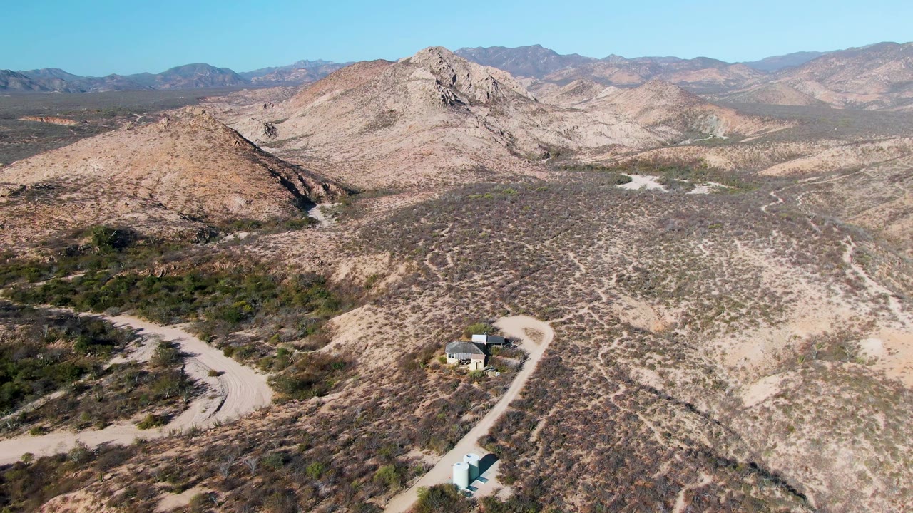 Cabo Pulmo aerial view with a small structure near the lower edge, pale dirt tracks through scrubland, rocky hills in the midground, distant mountains, clear sky, and no visible people.