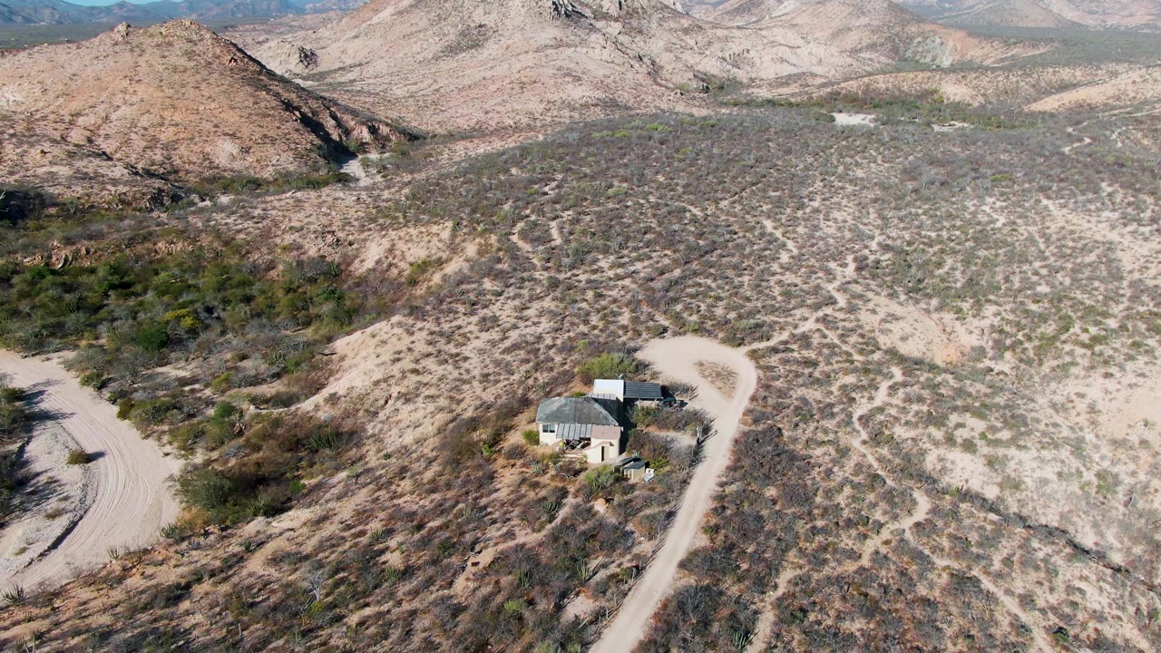 Wide aerial frame over Cabo Pulmo desert terrain with a curving dirt road, a small house below center, tan hills beyond, and no visible people.