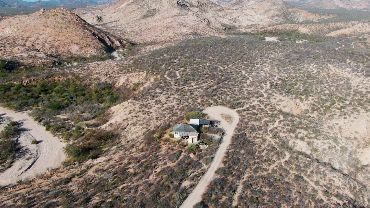 High aerial view of scrubland, a small house, a curving dirt road, and hills.