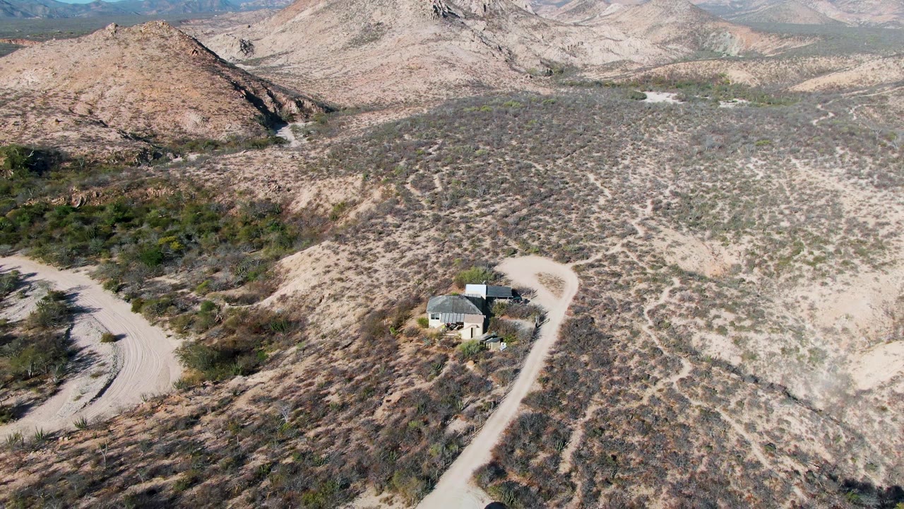Cabo Pulmo desert frame with a small structure near the road, scrub-covered ground across the foreground, layered hills in back, and no visible people.