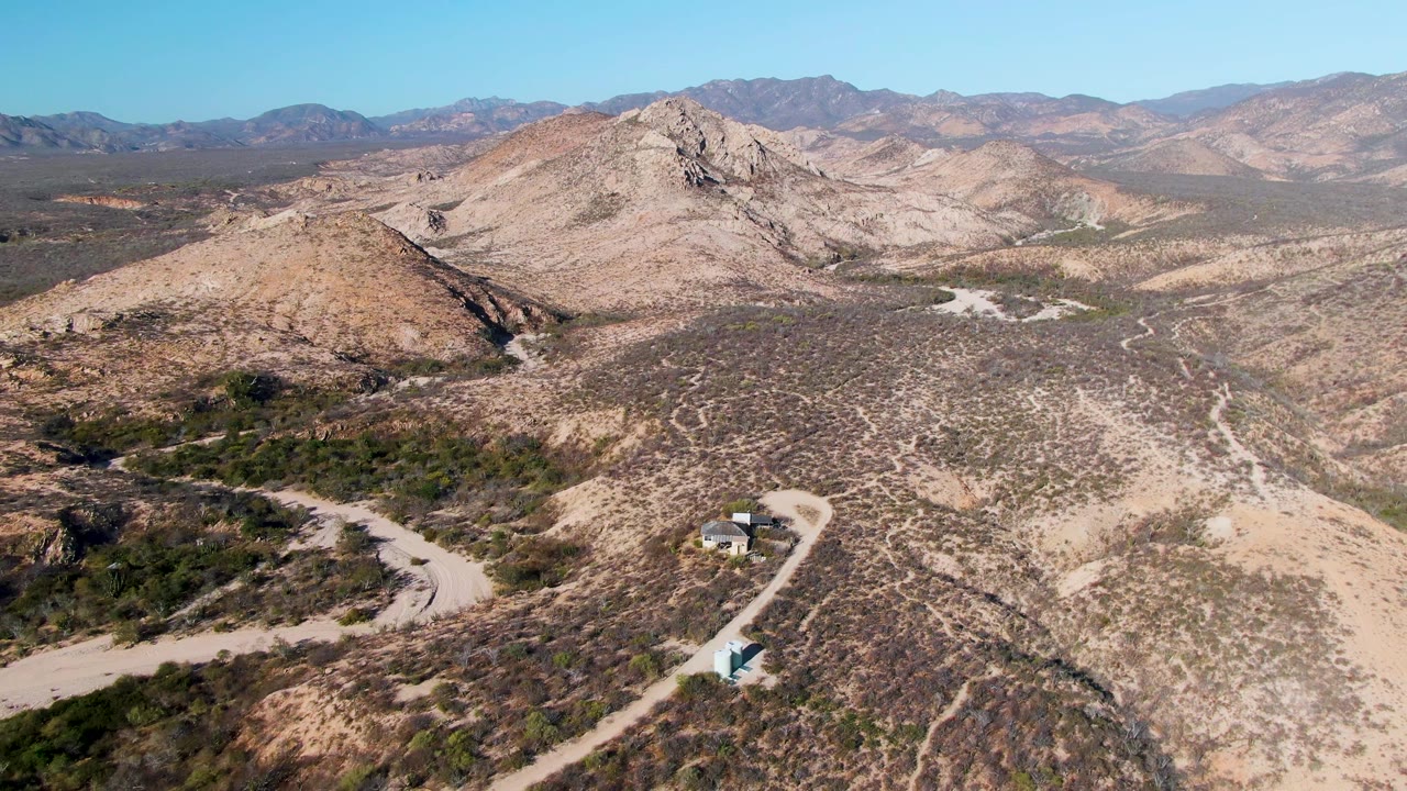Aerial view in Cabo Pulmo, Baja California Sur, showing the vehicle slightly farther along the pale road, with scrub-covered slopes, a rocky central hill, mountain ranges, and no visible people.