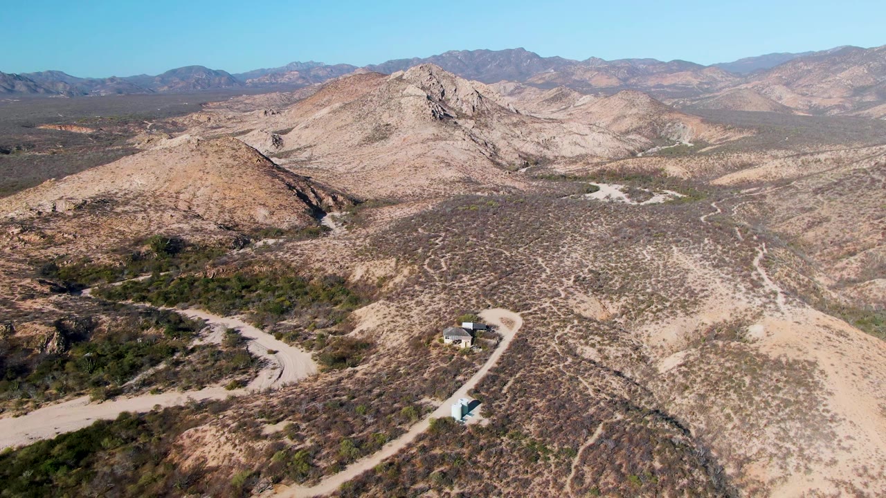 Drone-style aerial frame over Cabo Pulmo, Baja California Sur, with the road curving through tan scrubland, a small vehicle near the lower right road area, layered mountains, and no visible people.