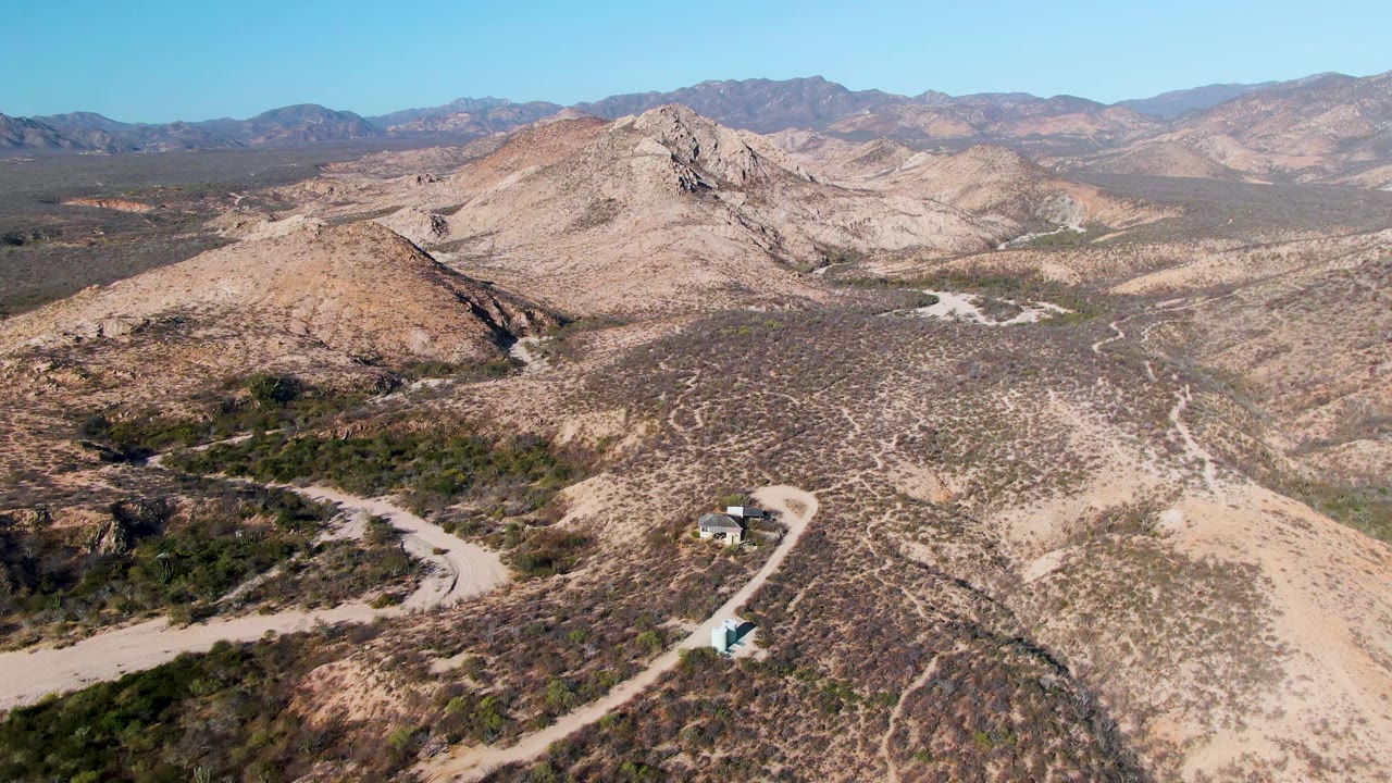 Wide aerial frame in Cabo Pulmo, Baja California Sur, with the vehicle visible on the lower winding road, a broad dry valley, rocky hill faces, distant mountains, and no visible people.