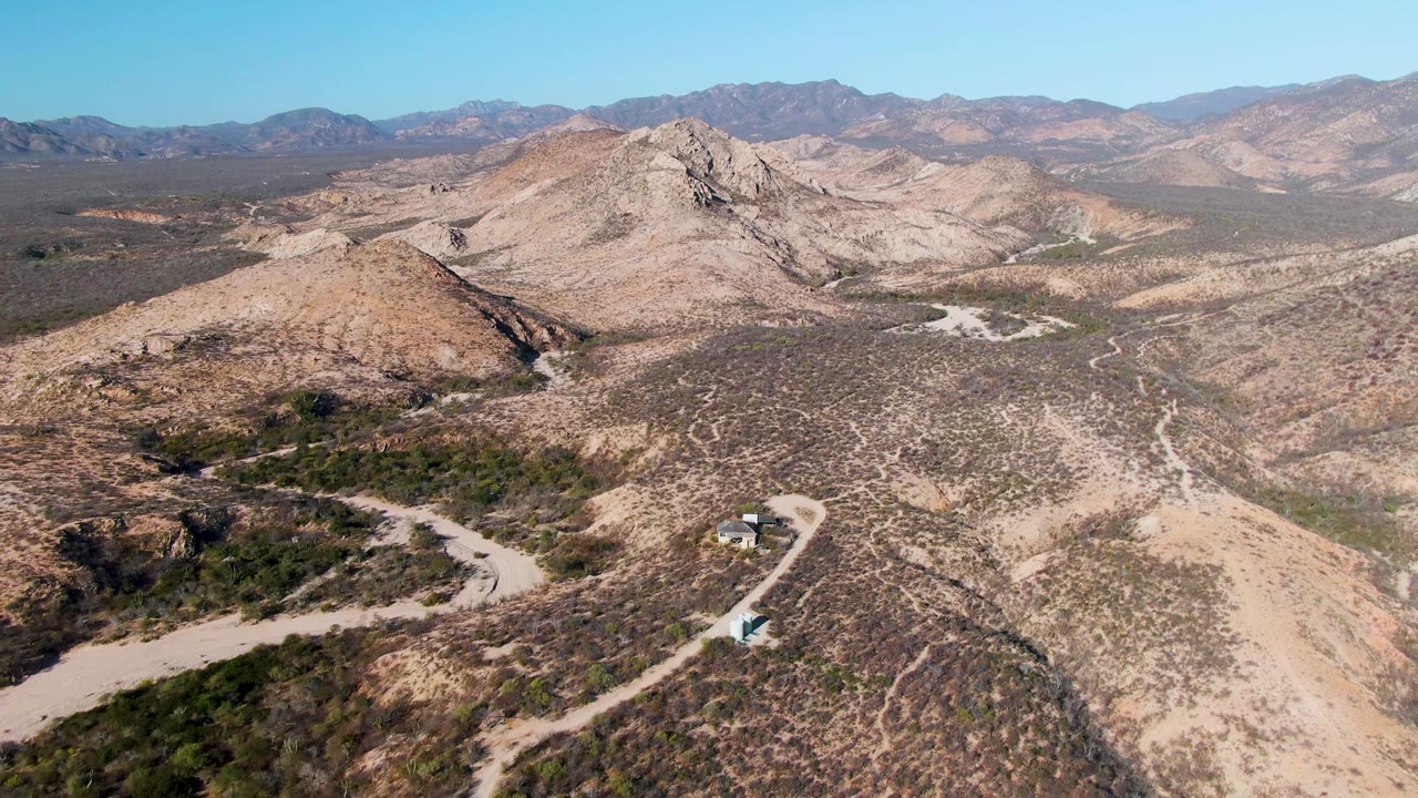 Wide aerial view over Cabo Pulmo desert hills with pale dirt roads curving through sparse vegetation, distant mountains, clear blue sky, and no visible people.