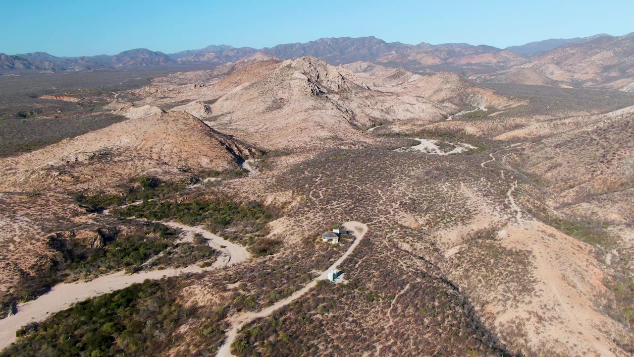 Cabo Pulmo aerial frame with a rocky hill centered, pale dirt tracks crossing the arid basin, layered mountains on the horizon, bright daylight, and no visible people.