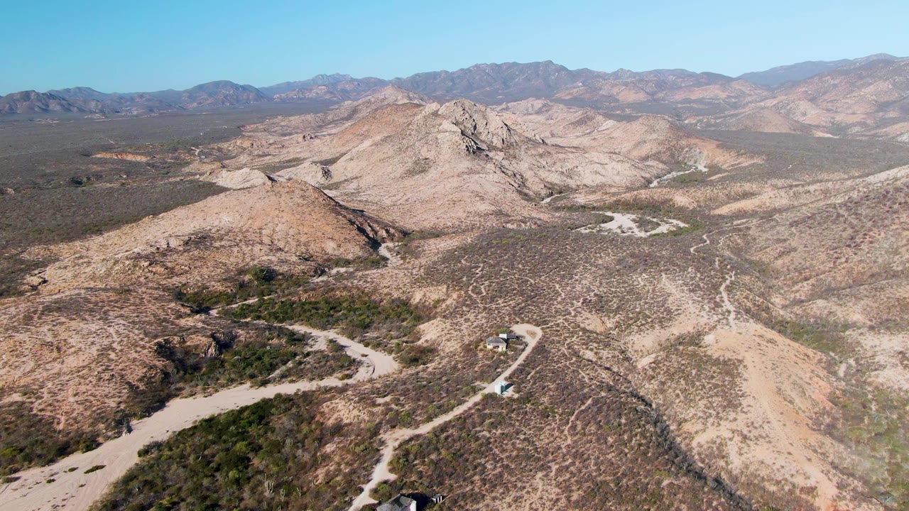 Aerial frame from Cabo Pulmo showing the vehicle slightly farther along the pale road, with scrubby slopes in front, tan ridges beyond, and no visible people.