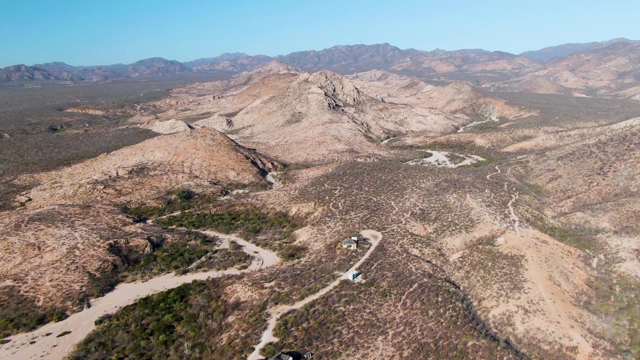 Elevated Cabo Pulmo view with the dirt road threading through the lower hills, the vehicle small near the bend, broad arid ridges, and no visible people.