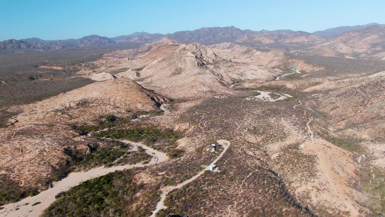 Cabo Pulmo desert frame with the road curve lower in the image, a small vehicle on the track, rocky tan slopes, distant mountain bands, and no visible people.