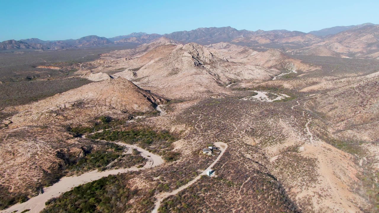 At 0:00, an aerial view near Cabo Pulmo shows a small vehicle on a pale dirt road below scrub-covered hills, with distant mountains and no visible people.