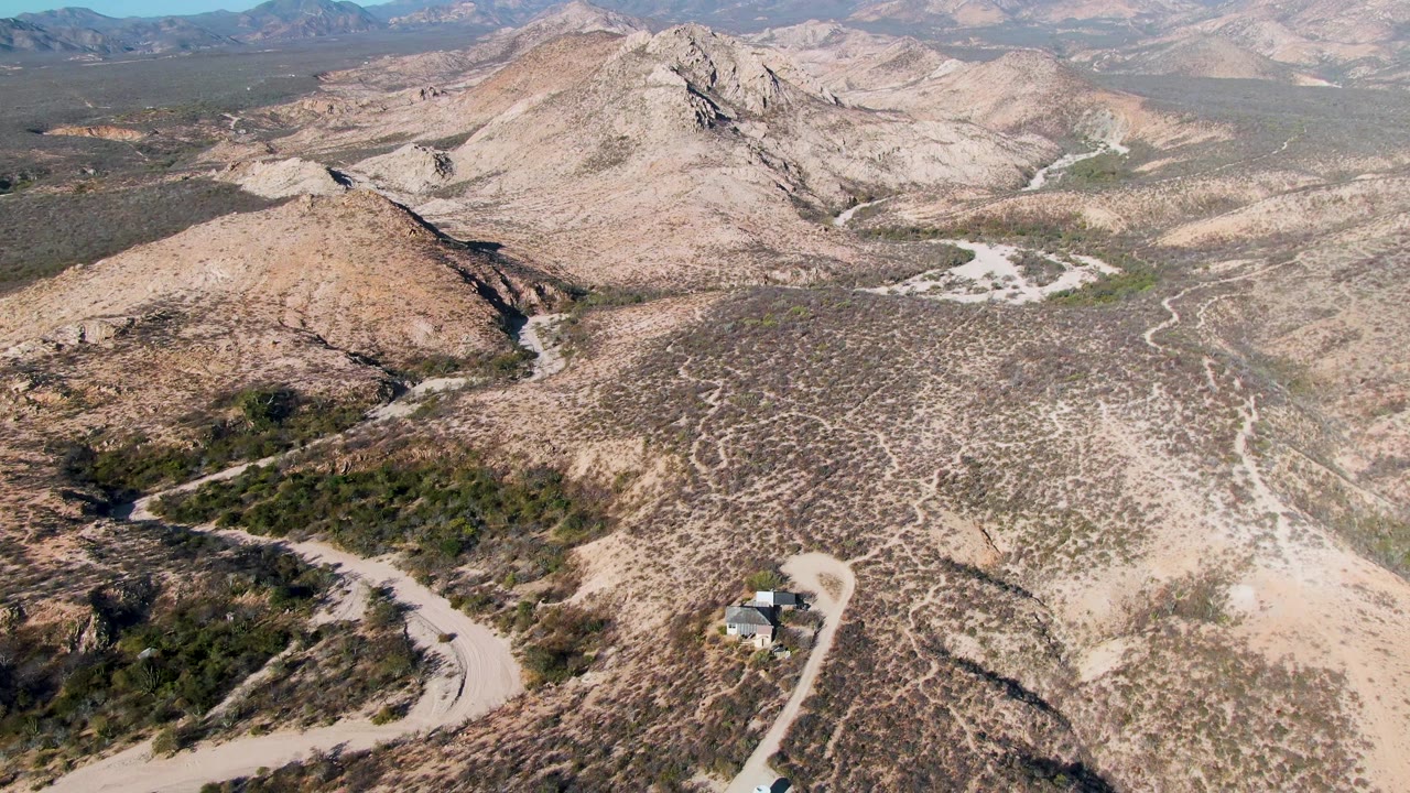 Aerial view over Cabo Pulmo desert hills with pale winding dirt roads, sparse scrub, distant mountains, and no visible people.
