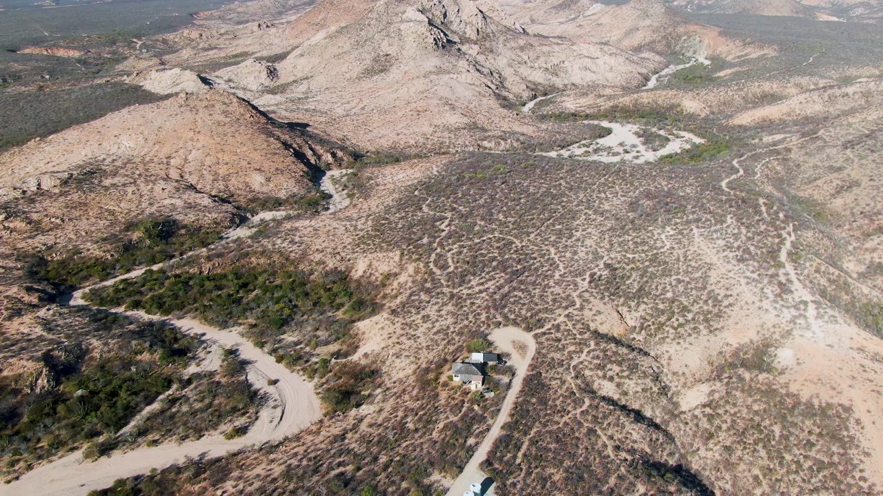 Drone view in Cabo Pulmo showing arid tan hills, a curving dirt road, a small roadside structure, sparse vegetation, and no visible people.