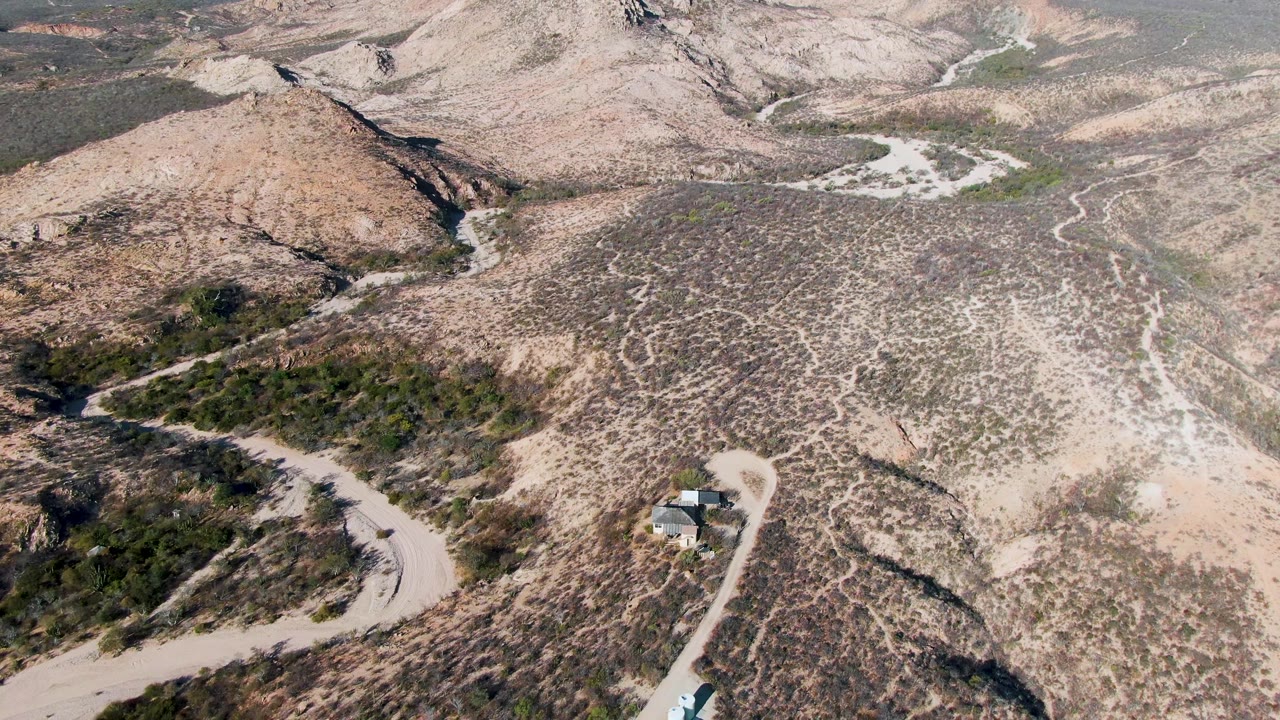 High aerial frame over Cabo Pulmo with pale tracks crossing dry scrubland, rocky slopes, distant ridgelines, and no visible people.