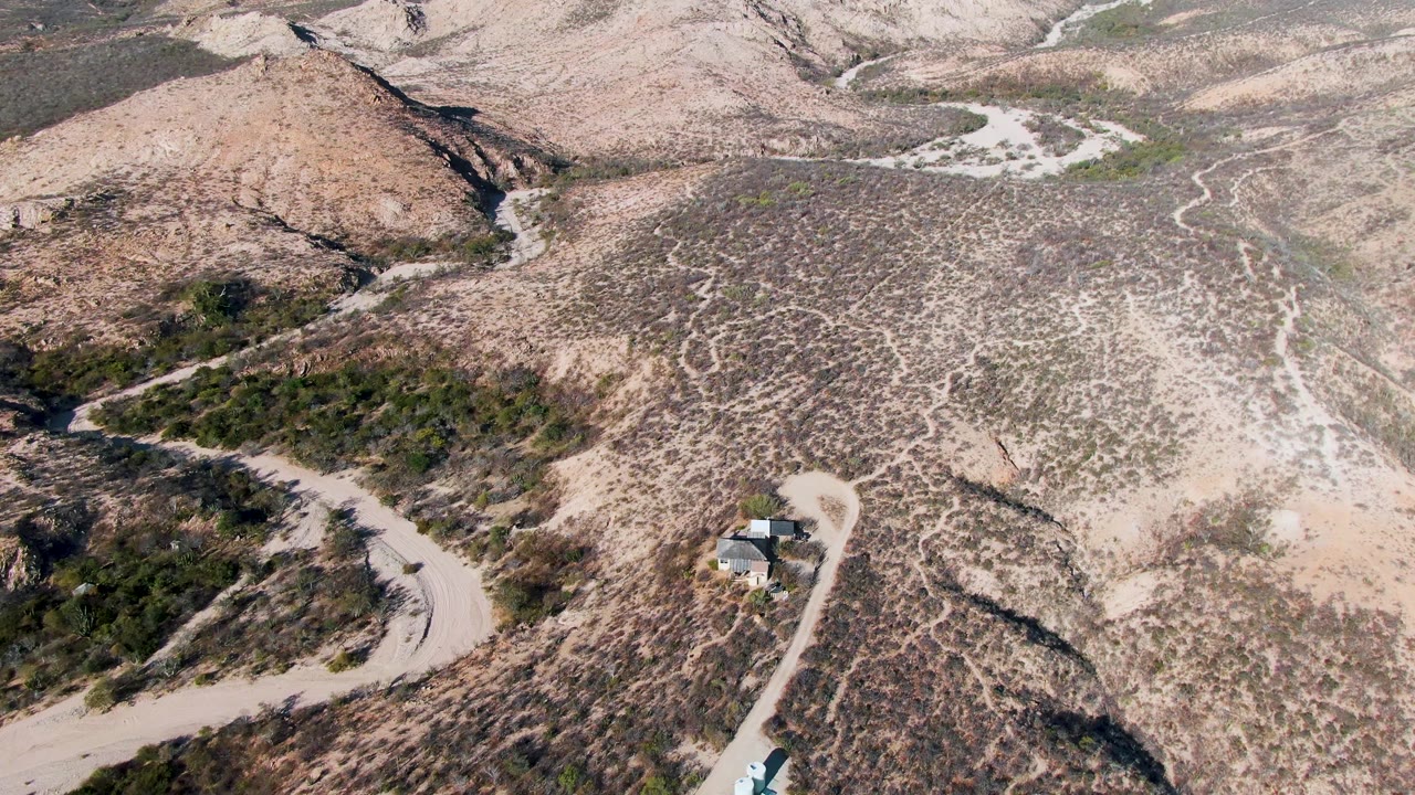 Aerial frame of a dirt road bending around scrub-covered desert hills.