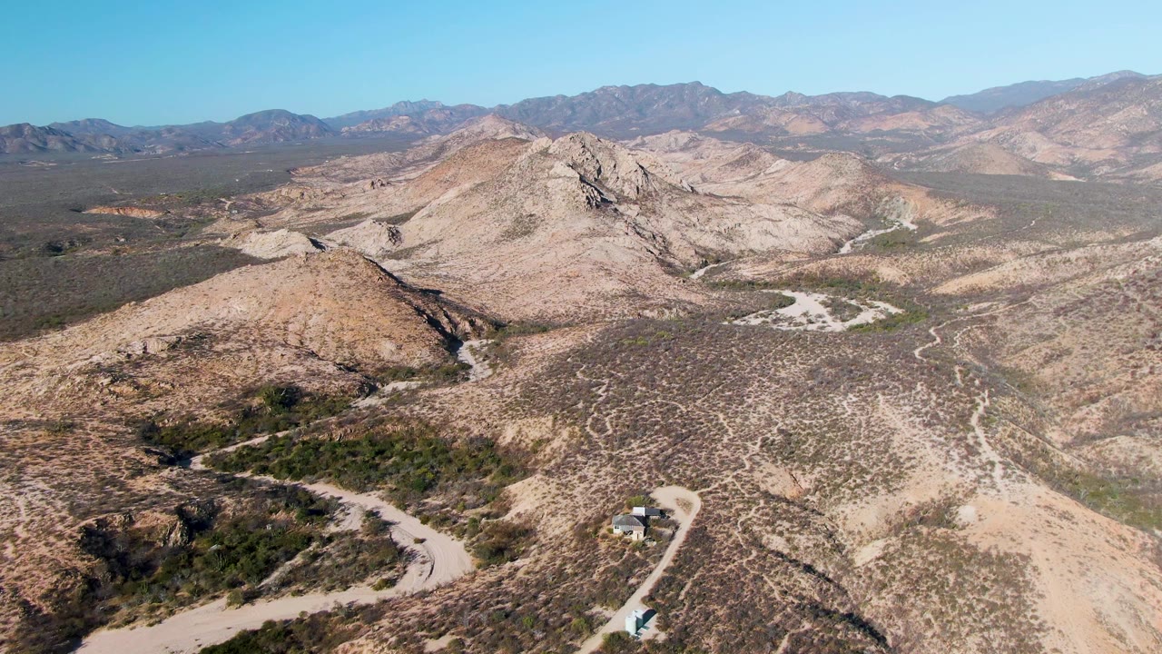 High aerial view near Cabo Pulmo, Baja California Sur, showing a pale dirt road curving through tan rocky hills, sparse desert scrub, distant mountains, and no visible people.