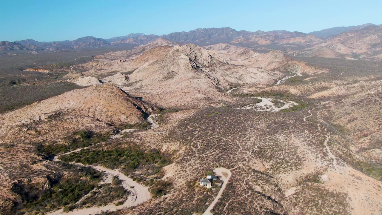 Wide aerial frame near Cabo Pulmo showing a winding road across arid slopes, a small vehicle near the lower edge, scrub-covered flats, distant mountains, and no visible people.