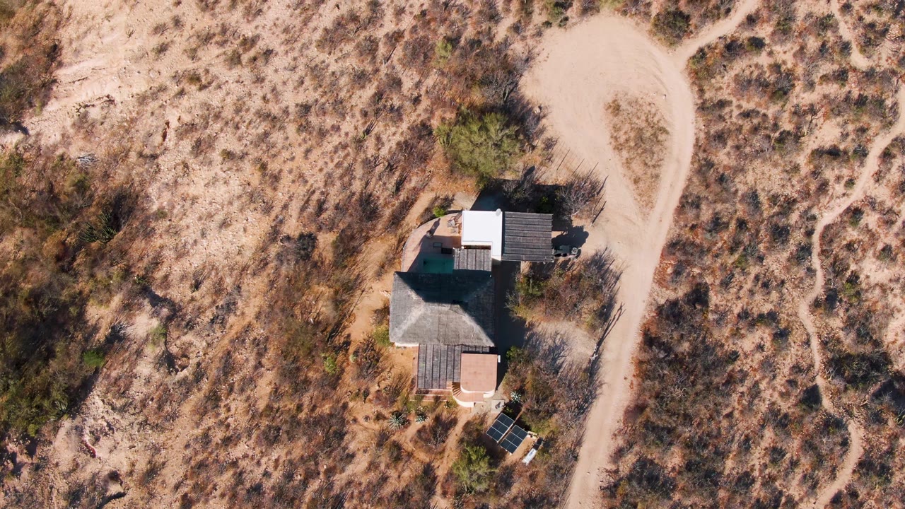 Overhead drone frame of a small house beside a pale winding dirt road in Cabo Pulmo, Baja California Sur, with dry scrubland, scattered green shrubs, hard shadows, and no visible people.