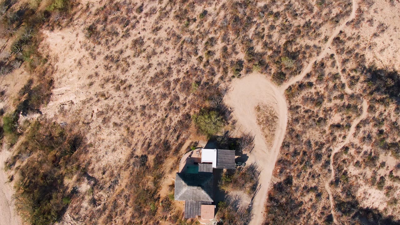 Aerial frame of the desert home near the lower right of the view in Cabo Pulmo, Baja California Sur, with a looped pale dirt road, scrub-covered slopes, and no visible people.