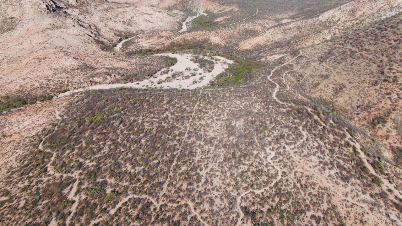 High aerial view over rocky desert hills near Cabo Pulmo, Mexico, with pale dry washes, sparse scrub, distant ridges, and no visible people.