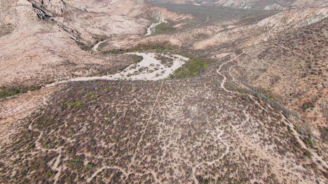 Aerial frame near Cabo Pulmo, Mexico, showing a winding dirt road through tan desert hills, dry wash channels, distant mountains, and no visible people.