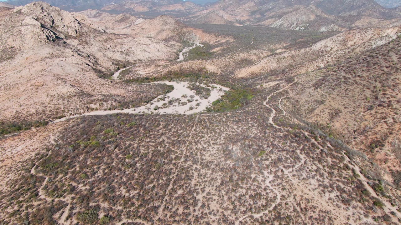 Wide drone view near Cabo Pulmo, Mexico, with a pale dry wash cutting across arid hills, scrub-covered slopes, layered mountain ridges, and no visible people.