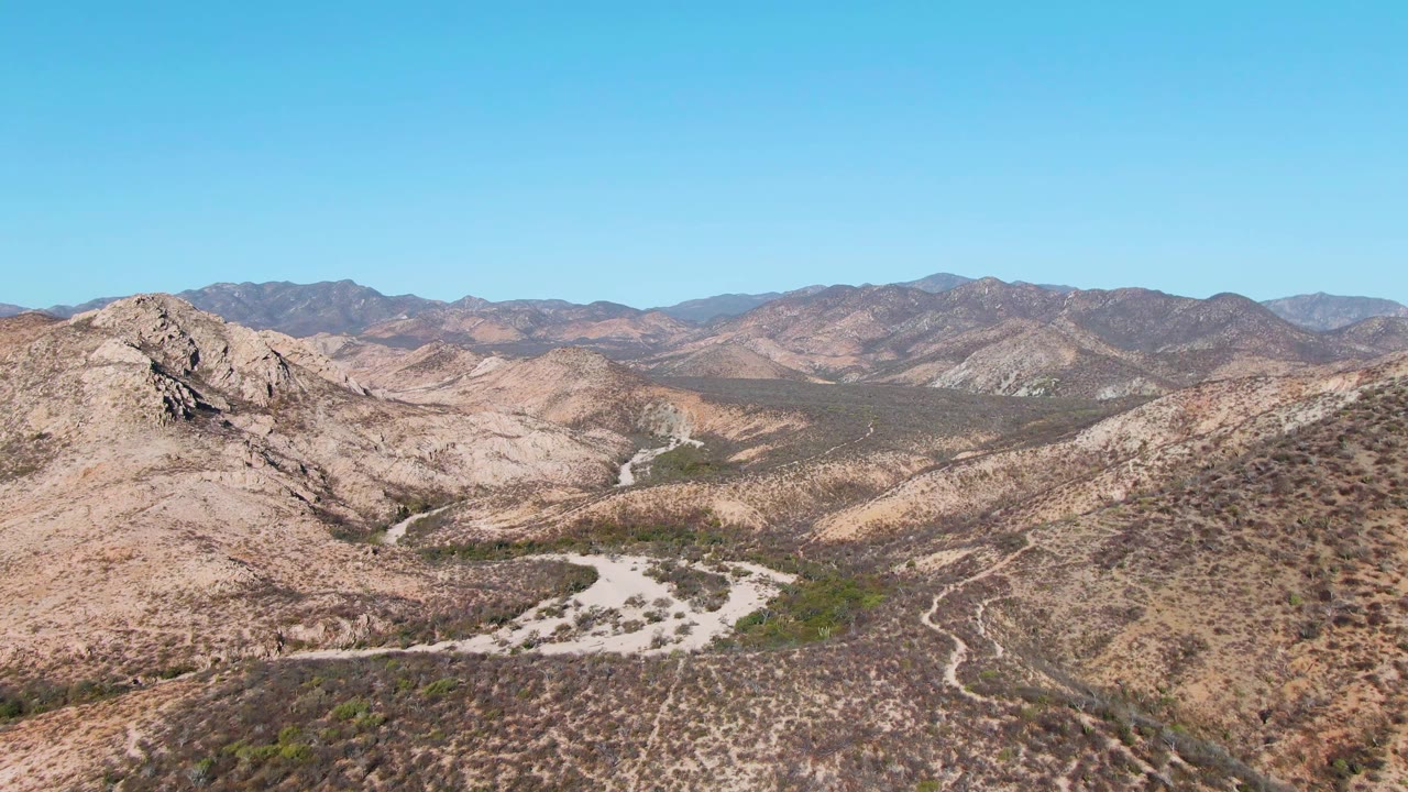 Second frame over Cabo Pulmo, Baja California Sur with the rocky ridge more central, tan slopes in the foreground, distant ridgelines, clear sky, and no visible people.