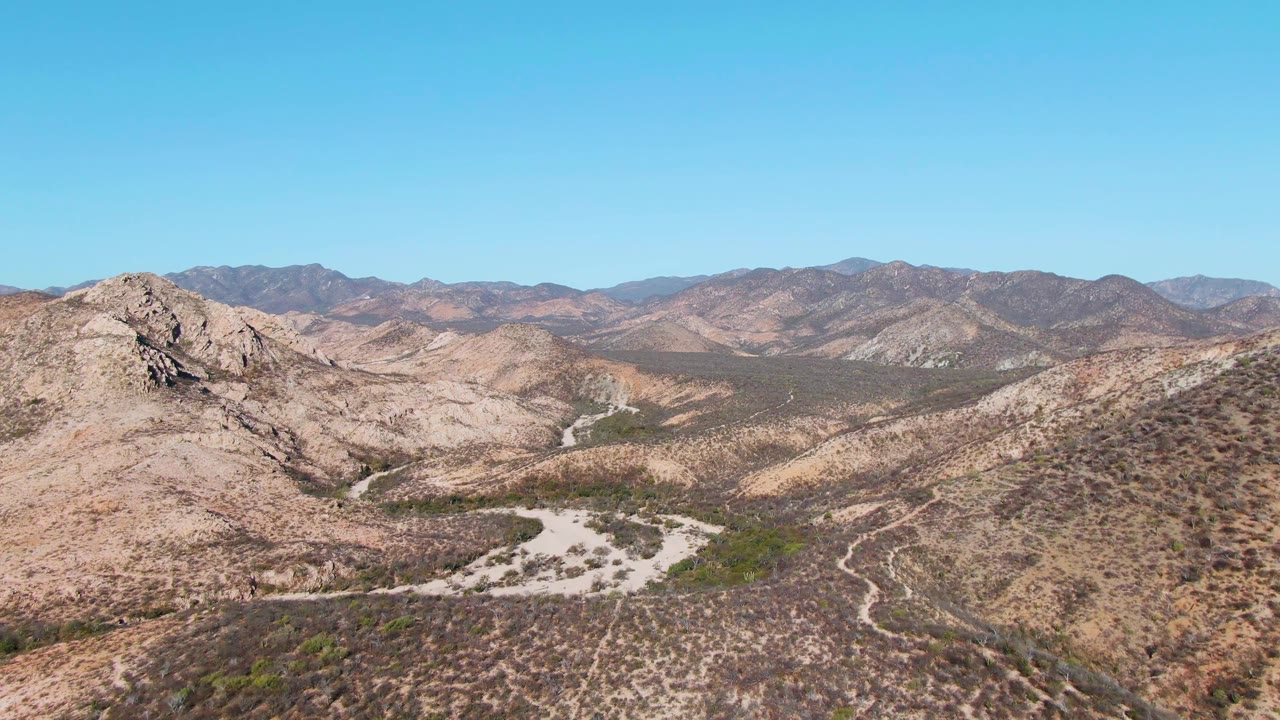 Third frame over Cabo Pulmo, Baja California Sur showing the pale winding road or wash between rocky hills, low desert vegetation, mountain backdrop, and no visible people.