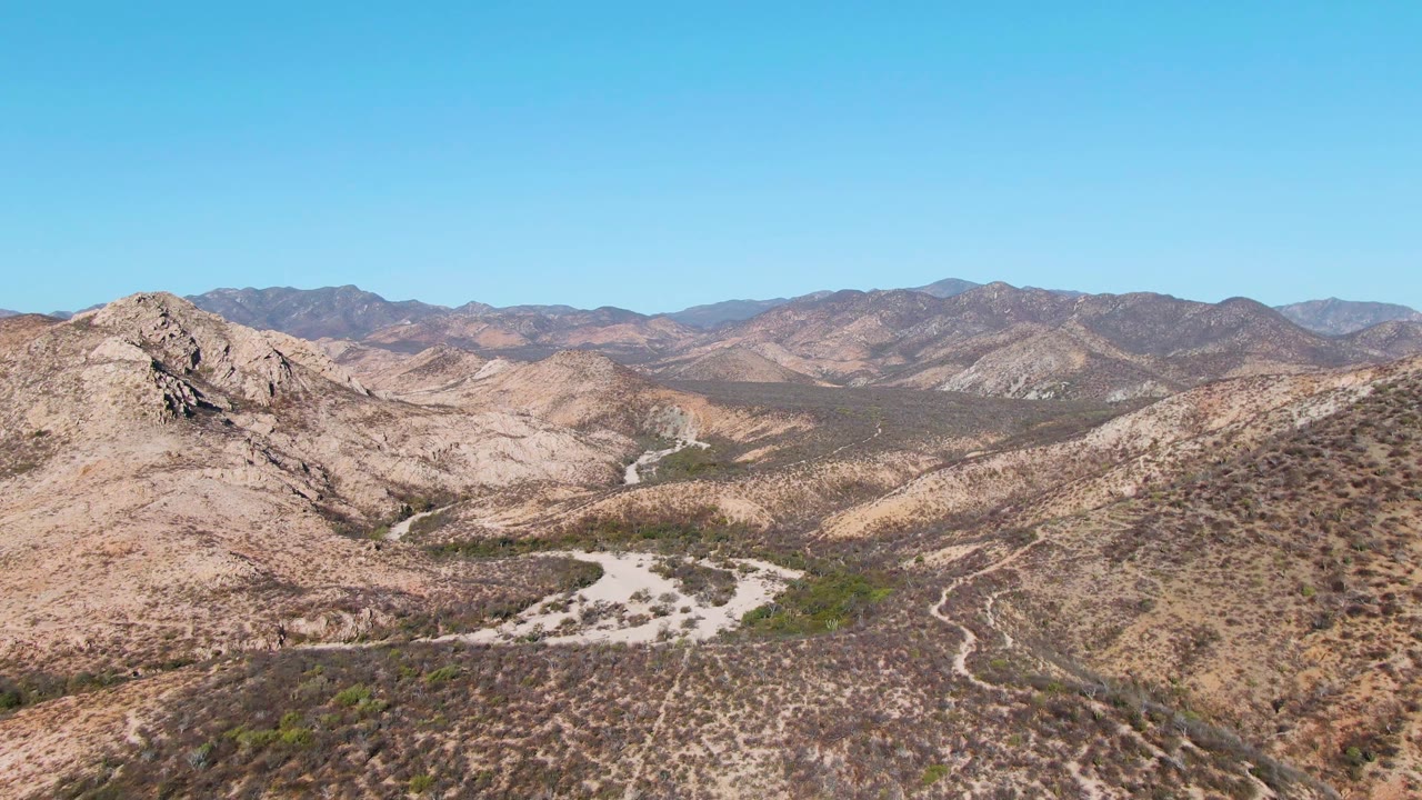 Fourth frame over Cabo Pulmo, Baja California Sur showing the view shifted across the rocky hills, with sparse vegetation in the foreground, layered mountains beyond, and no visible people.