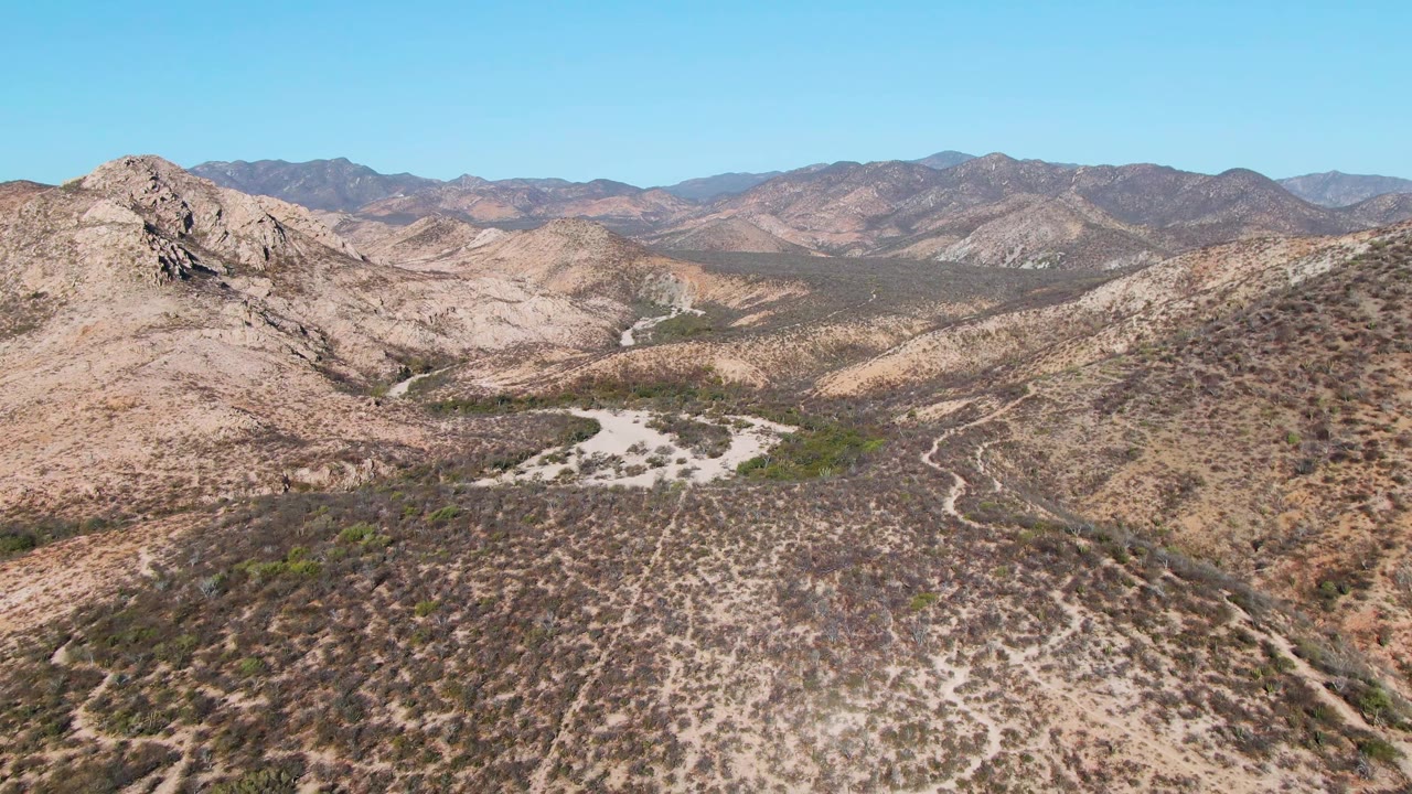 Opening aerial frame over Cabo Pulmo desert hills with sparse scrub, a rocky peak on the left, distant mountain ridges, clear blue sky, and no visible people.
