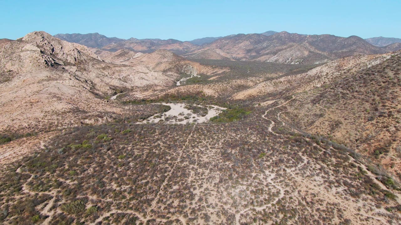 Aerial frame over Cabo Pulmo's arid valley with tan ridgelines, dark scrub patches, a pale wash near the center, layered mountains, and no visible people.