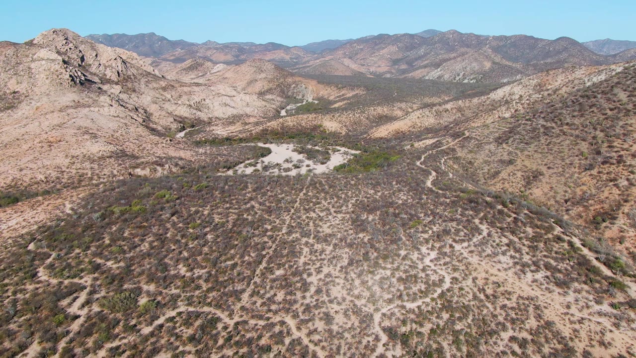 Aerial view in Cabo Pulmo showing a rocky peak left of center, dry rolling hills, winding dirt tracks on the right slope, distant ranges, and no visible people.