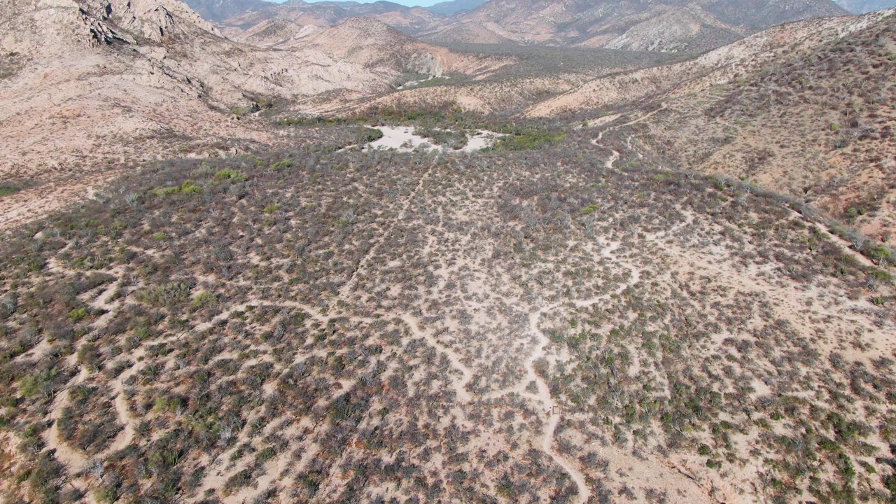 Wide aerial landscape of dry Baja hills with scrub patterns and a mountain horizon.