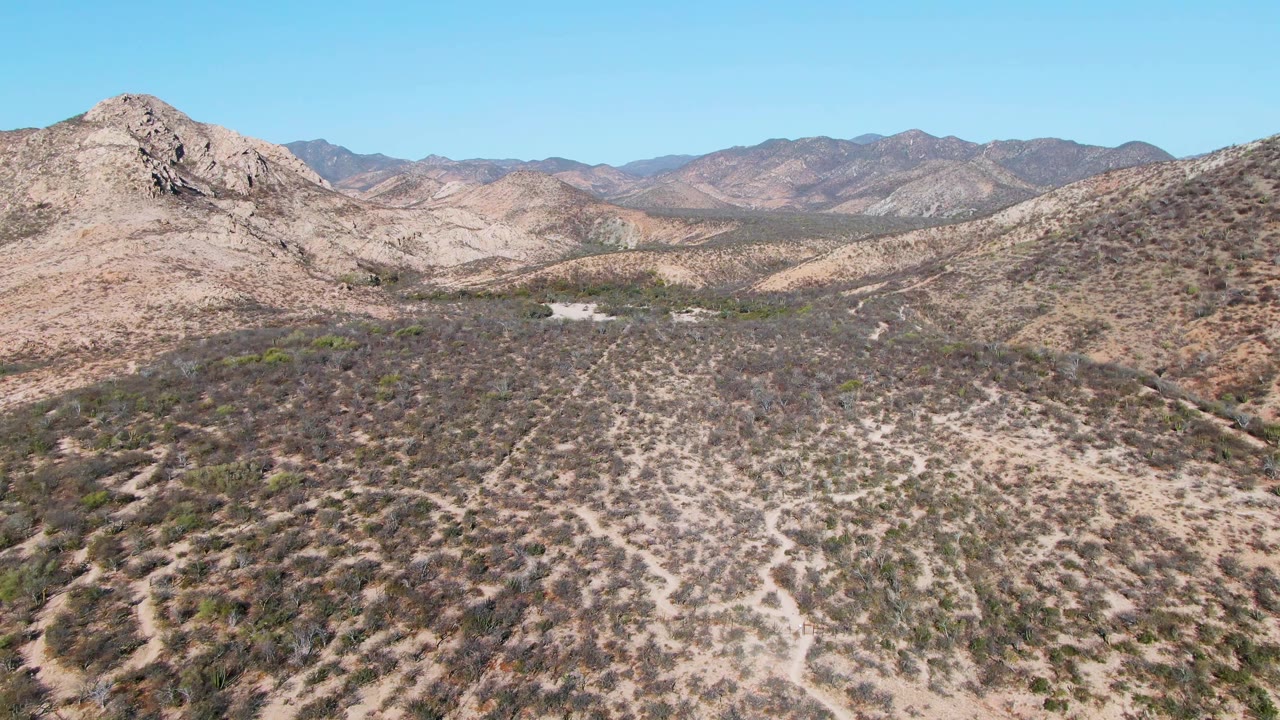 Cabo Pulmo aerial frame with a rounded rocky hill in the midground, patchy green shrubs on tan soil, distant mountains, and no visible people.