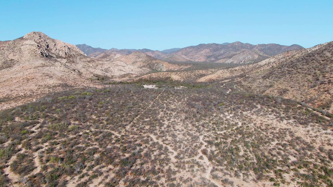 Mid-clip aerial view over Cabo Pulmo arid terrain, with low scrub in the foreground, a pale road or wash across the valley, mountain ridges, and no visible people.
