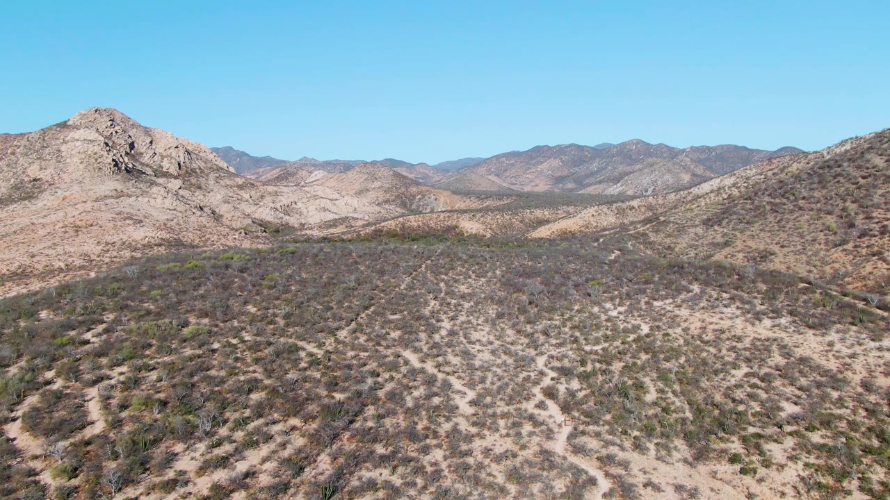 Wide aerial frame of the Cabo Pulmo desert valley with scrub-covered hills, pale dirt channels, distant rocky mountains, clear blue sky, and no visible people at the opening moment.