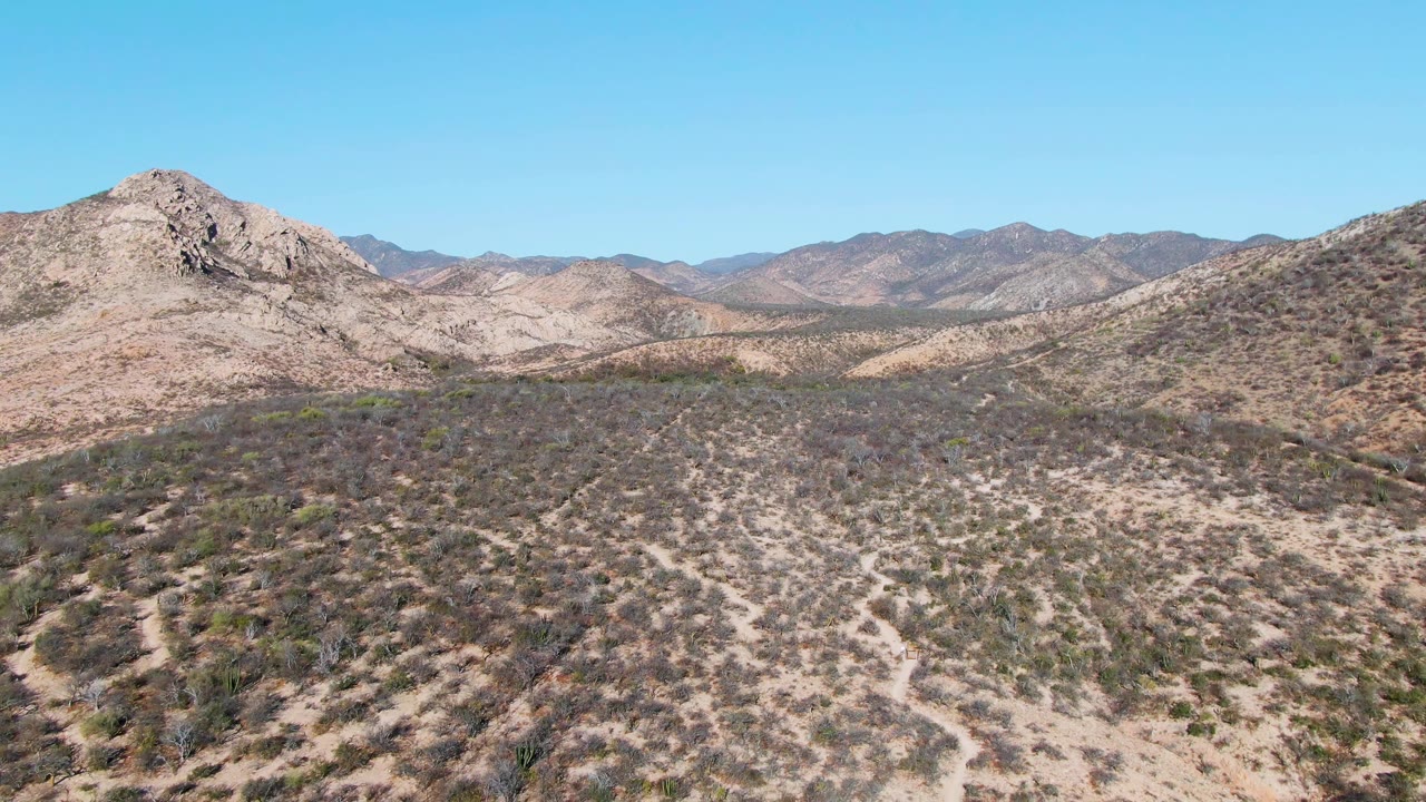 Dry valley with scrub, pale terrain, and rocky mountain ridges in the distance.