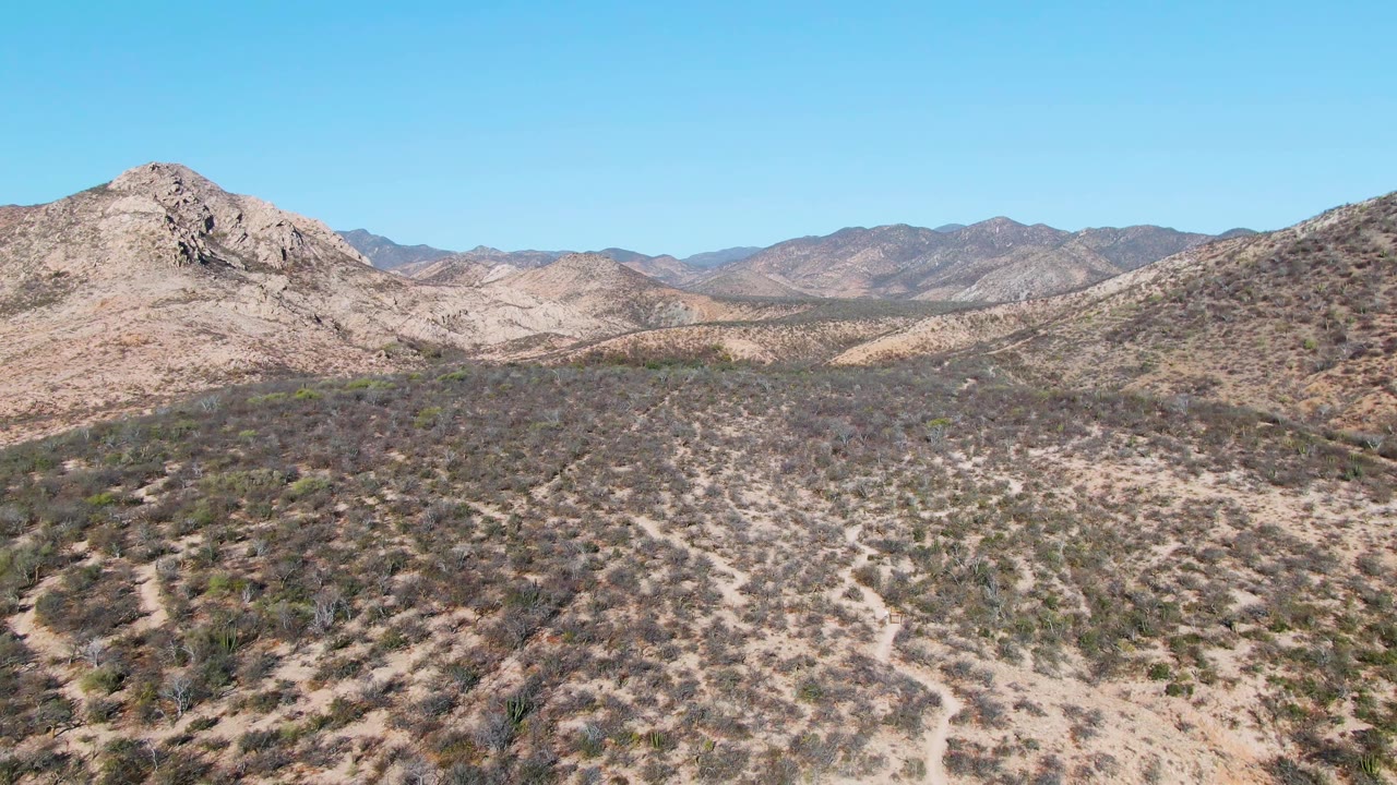 Mid-clip frame in Cabo Pulmo with a central rocky hill, dry scrub in the foreground, pale valley floor behind it, distant mountains, and no visible people.