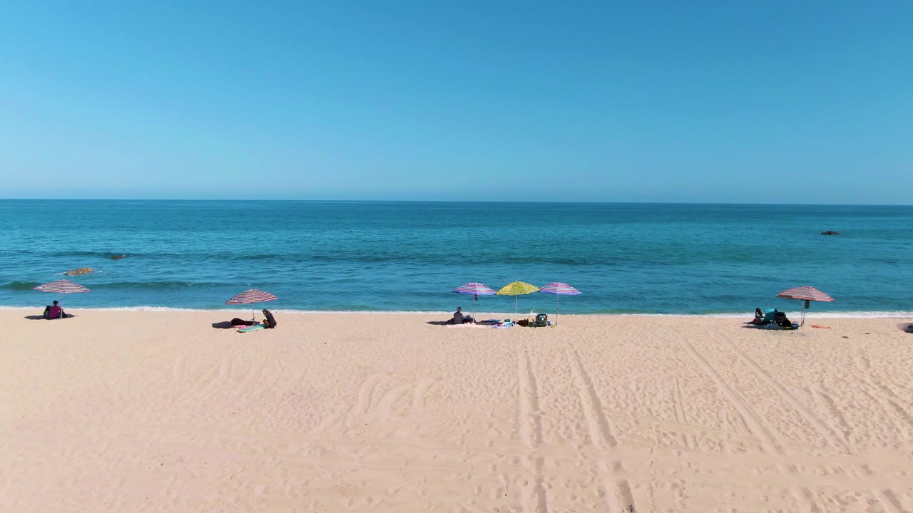 Aerial frame over La Fortuna beach shows scattered umbrellas and distant beachgoers on pale sand, with blue ocean and a clear horizon beyond.