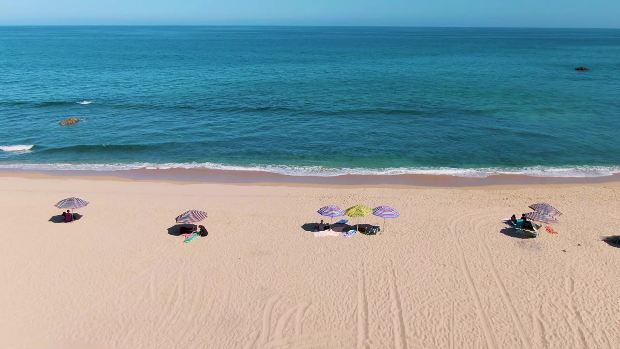 Aerial view of La Fortuna sand shows several umbrella groups spaced apart, with distant sunbathers below them and low white waves breaking along the beach.