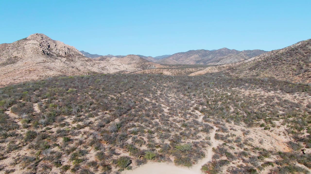 A wide aerial view in Cabo Pulmo shows a rocky central hill, low desert shrubs, layered background mountains, clear blue sky, and no visible people.