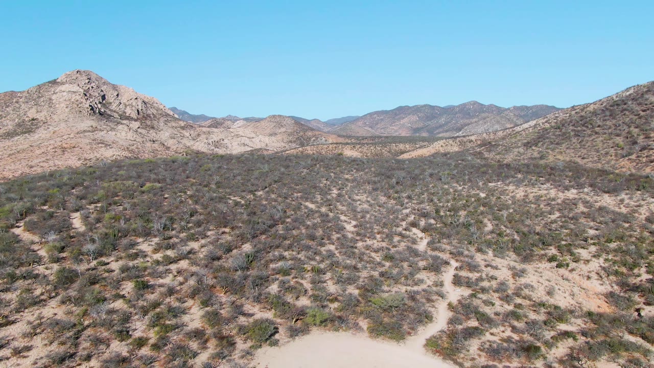 A Cabo Pulmo aerial frame shows scrubby desert hills and distant mountains while a pale dust trail spreads across the lower sandy foreground, with no visible people.