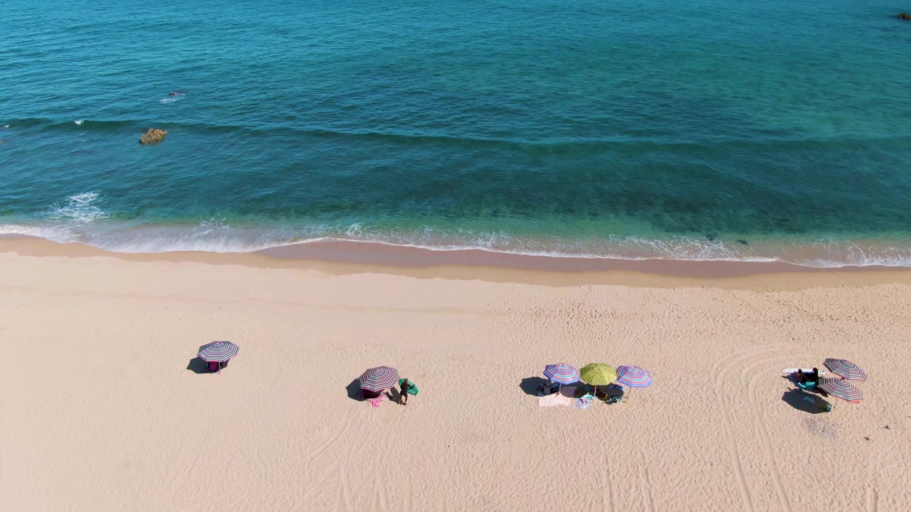 High aerial frame over La Fortuna beach in Baja California Sur, with pale sand, scattered umbrellas, chairs, tiny beachgoers, and blue-green water approaching the shore.