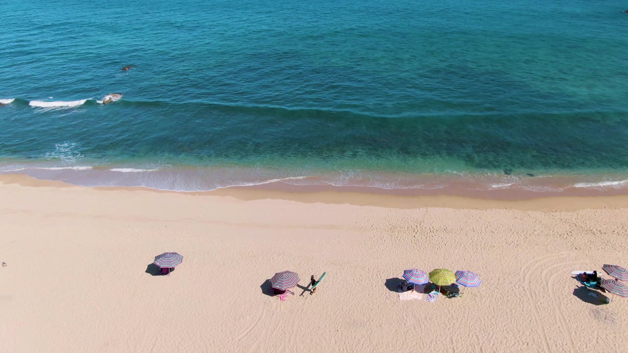 Top-down drone frame from La Fortuna showing a wide blue ocean band, pale shoreline, and several umbrella groups with chairs arranged on the sand.