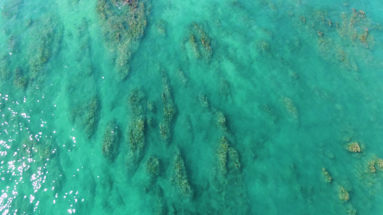 Aerial frame over La Fortuna coastal water showing dark green vegetation beneath turquoise water, pale sandy areas, and clustered sun reflections, with no visible people.