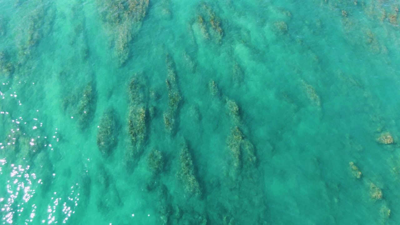 Clear turquoise water off La Fortuna shows elongated darker vegetation bands below the surface, with bright reflection specks near the lower frame and no visible people.