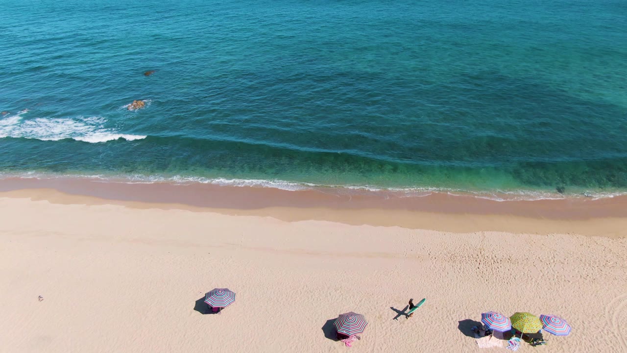 Top-down view of La Fortuna beach in Baja California Sur with several umbrellas on pale sand, turquoise water above them, small shore break, and no visible people.