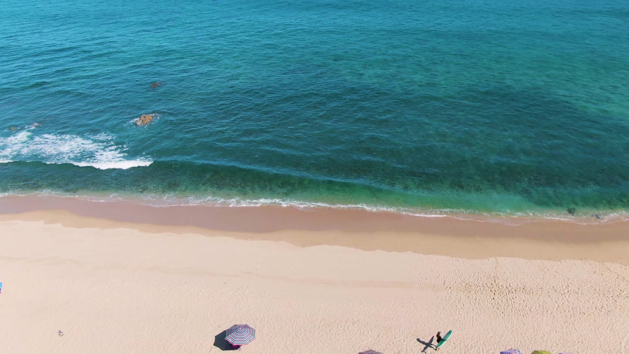 Aerial frame over La Fortuna showing a narrower band of sand with a few beach umbrellas near the bottom edge and blue-green ocean water rolling toward shore.